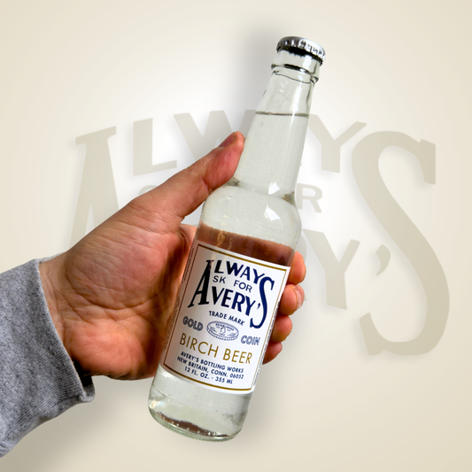 Hand holding a bottle of Avery’s Birch Beer in glass bottle on beige gradient background