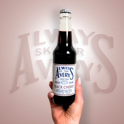 Hand holding a bottle of Avery’s Black Cherry Soda in glass bottle on deep red gradient background
