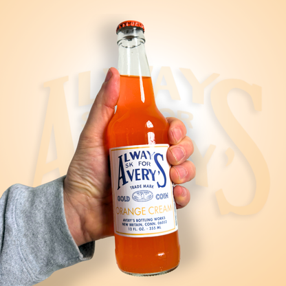 Hand holding a bottle of Avery’s Orange Cream Soda in glass bottle on orange gradient background